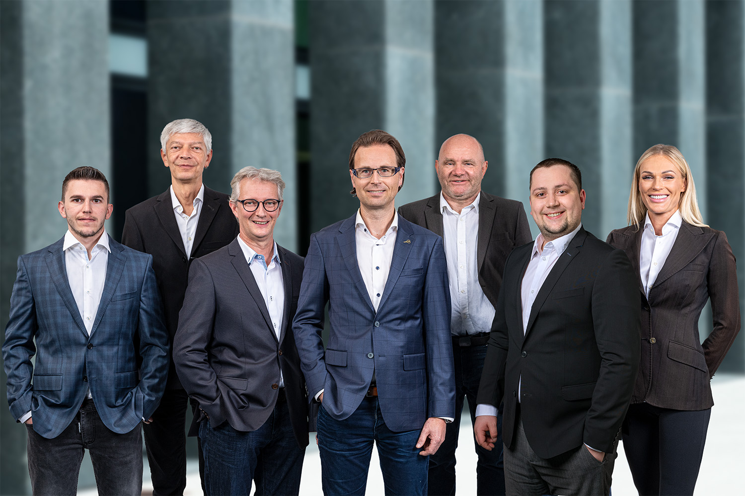 Sieben Mitglieder des Unternehmens Hemutec posieren für ein gemeinsames Teamfoto. In der ersten Reihe stehen vier Personen, darunter eine Frau, und in der zweiten Reihe drei Männer, alle lächelnd und in formeller Geschäftskleidung.
