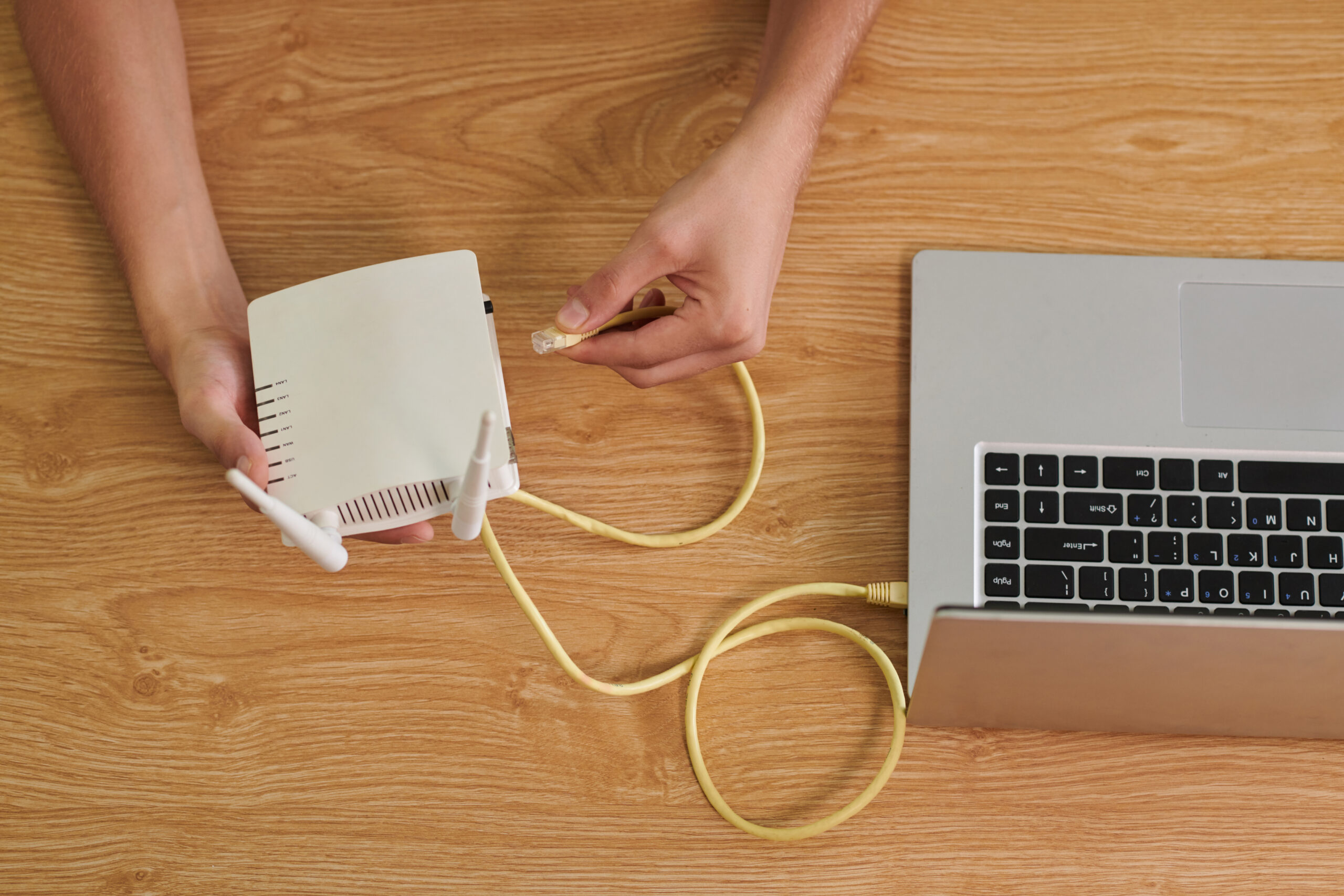 Eine Person schließt ein gelbes Ethernet-Kabel an einen weißen WLAN-Router an, der neben einem Laptop auf einem Holztisch liegt.